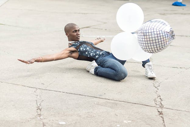 A performer supports their entire body with one bent leg to the ground as their upper body extends backwards, balloons are tied to them by a string on their neck.