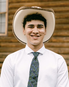 Portrait of Isai Soto in white collared shirt and blue paisley tie, he wears a tan cowboy hat in front of a wooden log wall