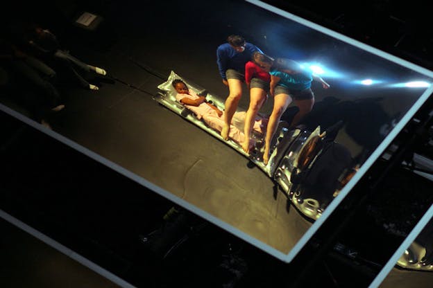 A photograph taken within a blue bordered mirror and slightly distorted. Three people wearing short grey shorts and primary colored t-shirts stand close together and balance, leaning forward over their front legs and concealing their faces. Behind them, a person wearing pink pajamas lays on a grey pool float.