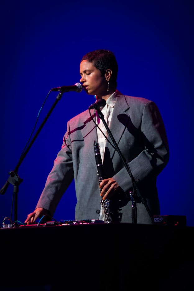 Holland Andrews stands against a blue background. One hand holds a clarinet and the other rests on a mixing board. In front of them are two microphones. Their eyes are closed and their head is faced downward. They are wearing a button down top with an unbuttoned grey textured blazer.