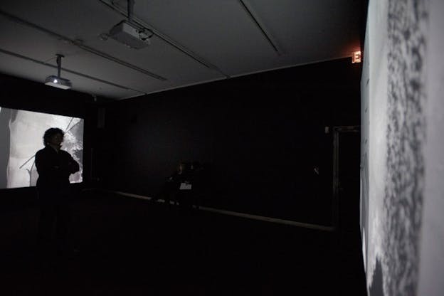 A black and white projection is shown in a dark installation space with one person standing in front of it.