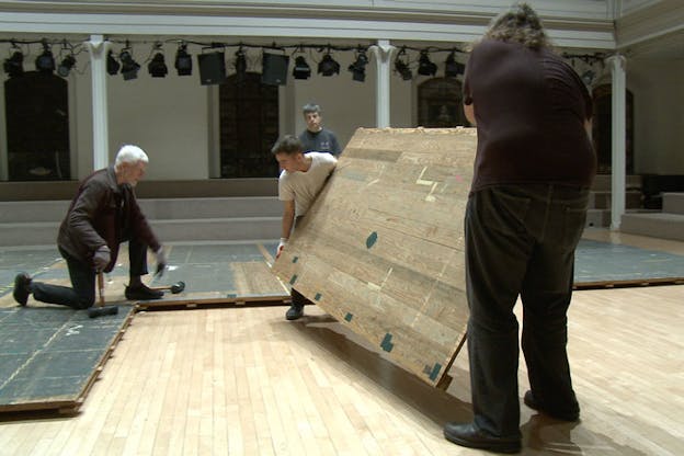 Two people hold a platform matching the other ones situated on the floor. A third person holding a hammer kneels in front of where the platform is to be situated. A fourth person watches from the back.  