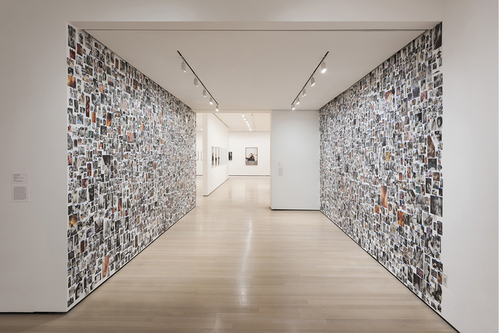 A gallery hall with beige wooden floors. On each side of the hallway collages of pictures cover the whole walls and deeper in the center at the end of the hallway, framed artworks. 