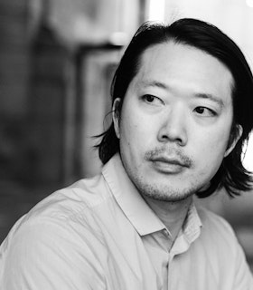 A black and white portrait of Kenneth Tam. He sits at an angle looking calmly off-camera over his right shoulder. He wears a collared button-down shirt.