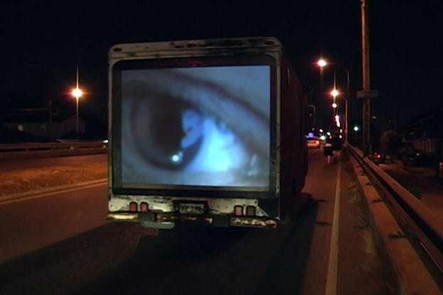 A photograph of the back of a truck on a street at night. A projection of a close up of someone's open eye plays on the back of the truck.