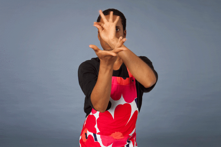 Harrell looks straight ahead, wearing a pink and white floral apron and twisting his hands over his face. 