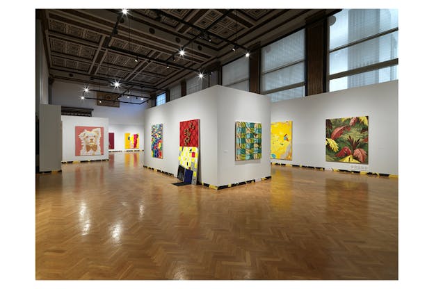 A gallery hall with various paintings. Some of them depict leaves of brown and green color, another depicts the portrait of a puppy and other more abstract ones in the color scheme of yellow, red and blue showcase different geometrical shapes in various fluid shapes.