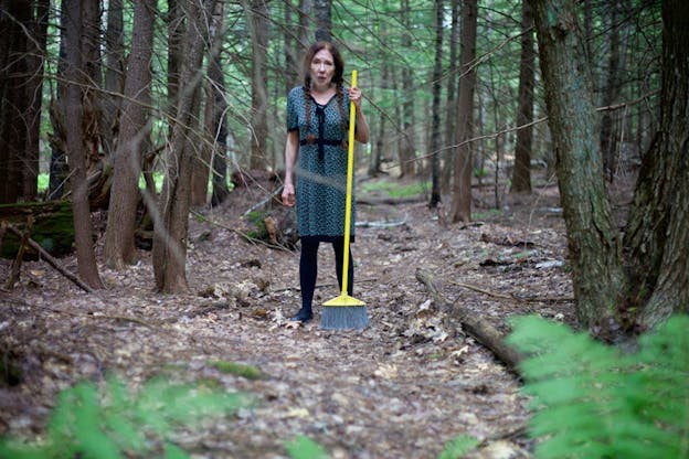 Performer stands holding a yellow broom in the center of bare trees, their mouth open and one eyebrow raised. Blurred green ferns line the front bottom and in the background, trees sprout green leaves. 