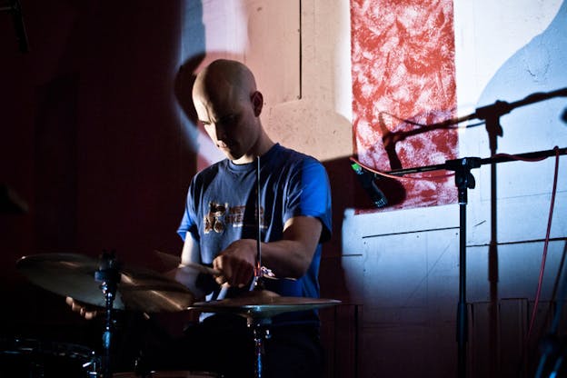 A figure playing the drums, behind them on the wall an illuminated area showcasing a red square. 