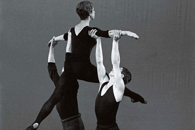 Black and white photograph of two people clad in black support another person's arms while they dance in the air.
