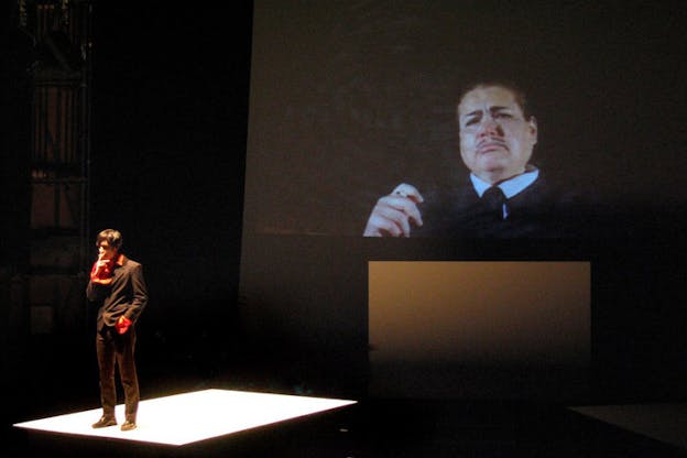 A figure standing in the middle of the stage with a cigarette in their mouth while a strong light centers on them. Behind them projected a man with a mustache, holding a cigar and frowning.