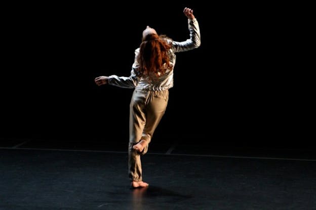 Performer facing away, tilting back their head,bending one knee, and holding their arms above and to the side in right-angles on a black-lit stage.