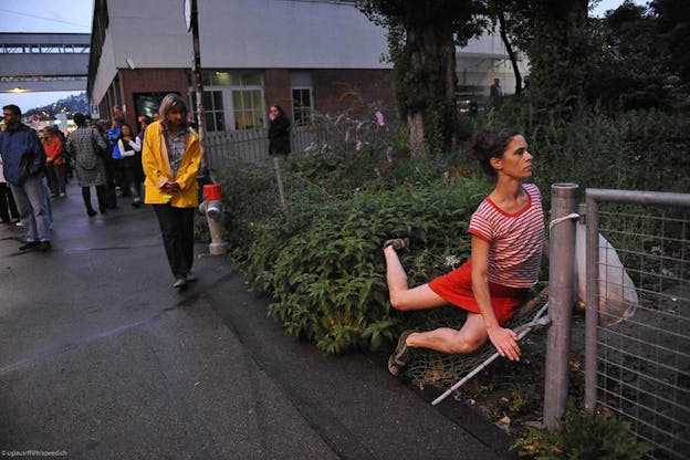 A person in a yellow coat, separate from a crowd, observes a person in red stripes kneeling and facing forwards with one back leg outstretched against a broken wire fence and an overgrown bush. 