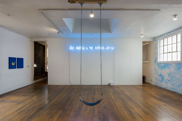 Image of a gallery room with a wooden floorboard and white walls. In its center a blue swing with metallic links hangs from the ceiling, behind it on the wall blue neon letter reading we deserve to see ourselves elevated.
