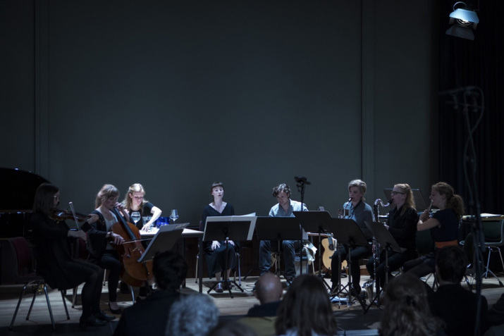 Image of an orchestra seated in a semi circle behind music sheet panels facing the audience, each holding a different instrument.