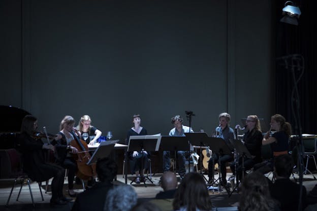 Image of an orchestra seated in a semi circle behind music sheet panels facing the audience, each holding a different instrument.