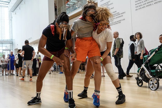 Three performers dressed in shorts and eye masks hold on to each other pushing forward with their faces strained while people in line behind them watch.