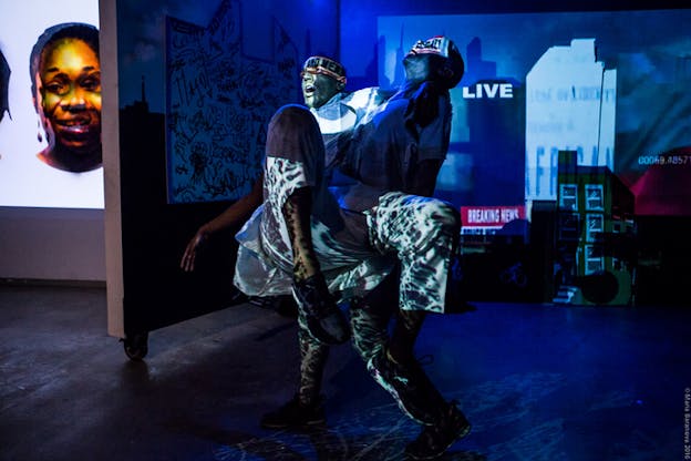 A performer lifts another one off the ground by holding under their arms and knees. Both are projected various shapes and words on them, while on their background is projected a collage of news titles and intros.