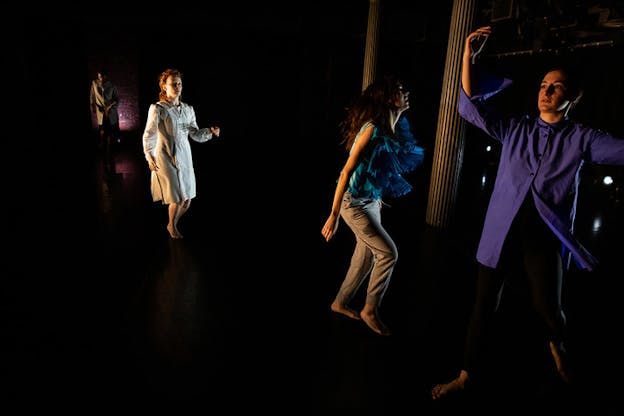 Performers on a dark stage stand, with their side towards the viewer, in a spacious line in various poses from left to right. On the end right closest to the viewer stands the only performer turned en face, in a movement of lifting both their arms on air and one of their legs, making the purple dress shirt move. 