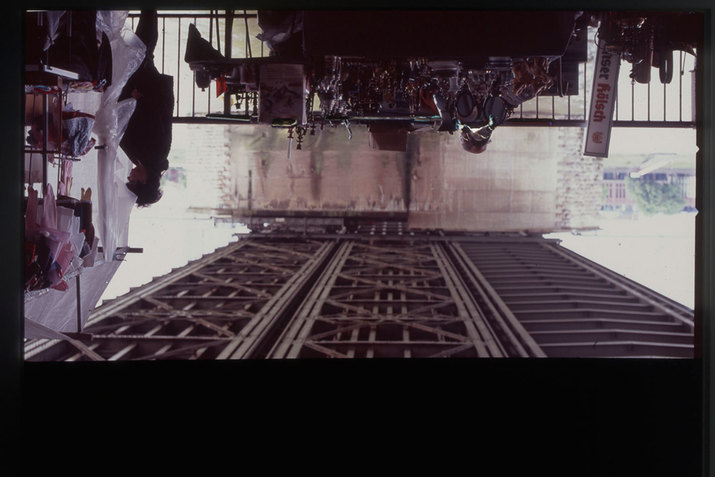  View of the criscrossed metal bottom of a bridge rising upwards while a table covered in shadowed, shiny objects and a person standing beside plastic tarp attached to a rack of children's clothes hang upside down.