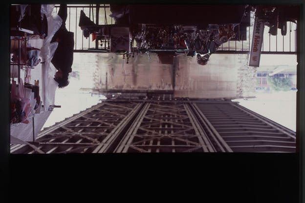  View of the criscrossed metal bottom of a bridge rising upwards while a table covered in shadowed, shiny objects and a person standing beside plastic tarp attached to a rack of children's clothes hang upside down.