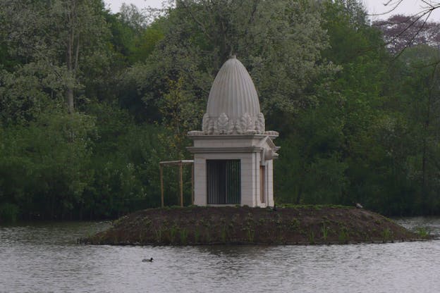 An image of Lincoln's Pig Palace situated on a small island in the middle of a lake surrounded by lush trees. The palace is made of entirely pale grey stone and has a square base with a pointed dome atop that. The dome is decorated with thick leaf-like carvings and the front of the square base has a pig head above the door. The base also has two tall black striped windows on opposite walls.