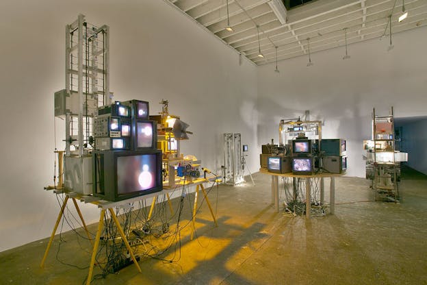 Several sculptures are situated within an otherwise bare white room. In the foreground, there is a sculpture comprised of a table with multiple different sized television screens on it all projecting a blurred image of a purple and blue hued light. In the background, there is another table of screens which project different blurred images. A metal ladder beside this table supports more screens.