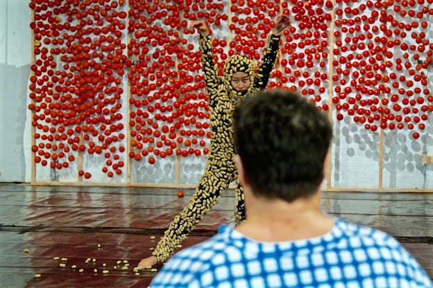 A performer dressed from head to toe in a black costume with peanuts on it stands with their arms upwards in front of a white wall with red spheres as a person with their back to the viewer watches them. 