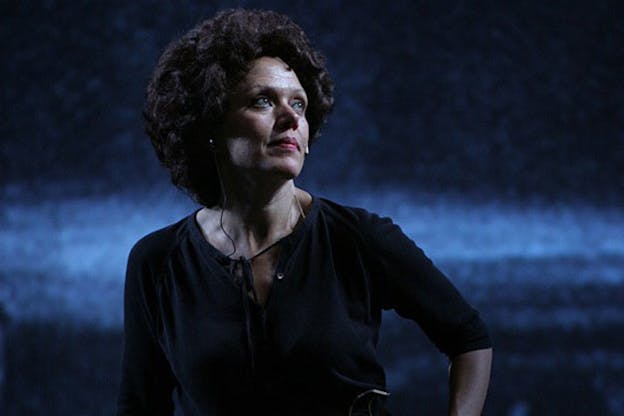 Kate Valk is in front of a dark blue background, wearing a black top. Her hair is short and curly. With her profile to the camera, she looks up wistfully to a point beyond the camera frame.