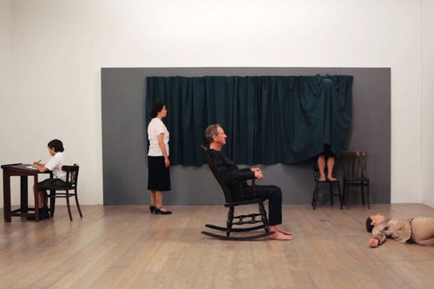 In the center of a room, a person sits on a sideways-facing rocking chair, on the right a person lies on the floor, in the left corner a child writes at a desk, and along the gray wall with a seaweed blue curtain, one person stands and faces the wall while another stands behind the curtain, only their feet elevated on a wooden chair visible. 