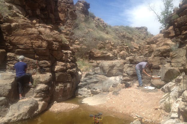 Two persons arrange recording instruments in the tannish-red rocks of a desert watering hole.