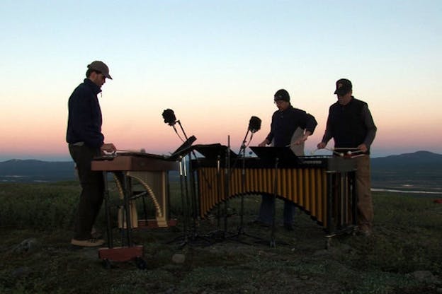 Three figures stand on a green grassy area playing marimbas connected to computers. Behind them the sky at the top is a sea blue and as it goes down to the horizon line, becomes a sunny yellow and a peachy orange that blend together. The view of the sky is cut by mountains with a river crossing in front of them.