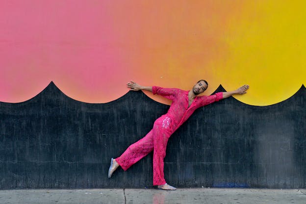 Mitchell wearing a hot pink jumpsuit faces forward and leans with his back leg raised and his arms outstretched against a gray wall with ombre pink, orange, and yellow sunset colored scallop print. 