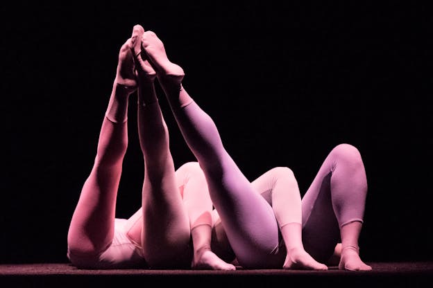 Three laid down aligned figures, with their bottom halfs only visible to the viewer lean their left leg on top of each other's right while their right legs are elevated and touching one another's at the toes.