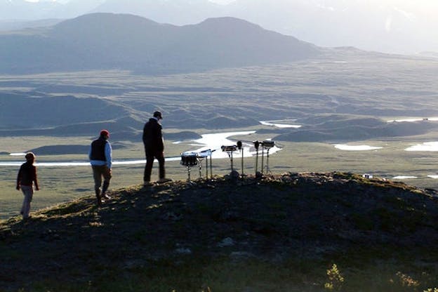 Three people walk in a queue upwards a hill towards three musical instruments. Behind the hillside there are grassy green plains with masses of glistering water and the end tail of a mountain.