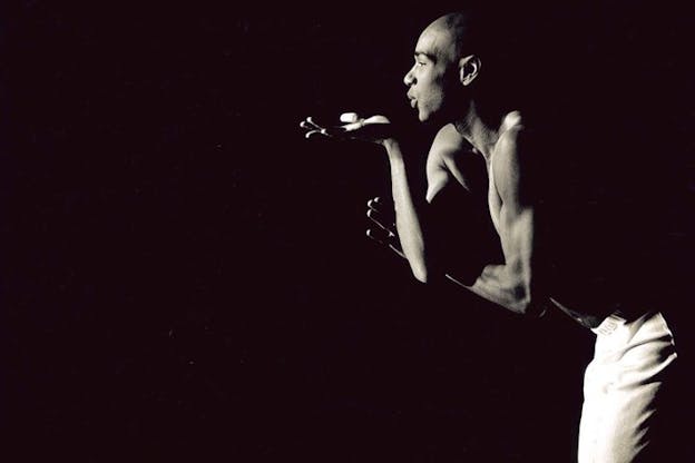 Black and white photograph of a shirtless figure on the side blowing a kiss with one hand palm facing upwards situated under their lips as the other loops underneath it.