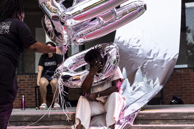 Autumn Knight sits on a sidewalk curb, holding her face in her hand. She wears a white linen outfit and holds a microphone in one hand. Next to her, someone holds out a collection of metallic ballons. One such balloon surrounds the performers head. A sheet made from the same metallic material hangs behind the performer. A bright pink light reflects on the performer and the balloons. A particially obscured audience member watches from behind.