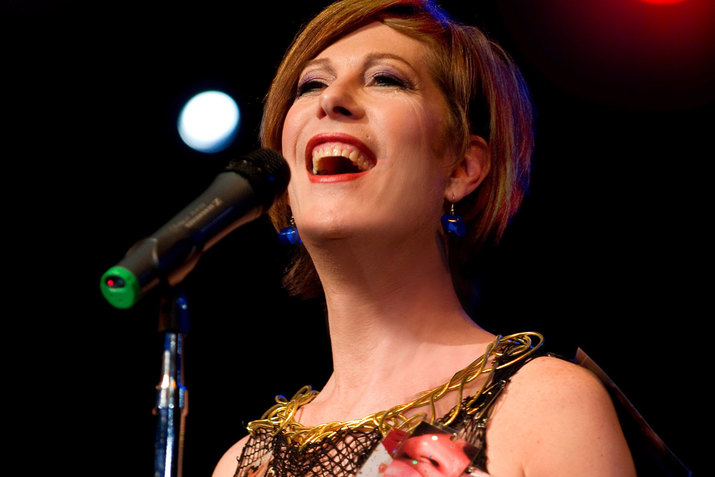 Low-angle close-up of Bond smiling into a microphone, wearing red lipstick, blue glass earrings, and a gold adorned top.