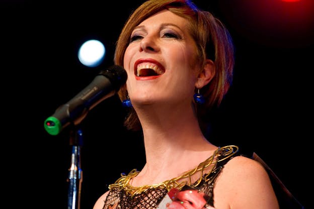 Low-angle close-up of Bond smiling into a microphone, wearing red lipstick, blue glass earrings, and a gold adorned top.