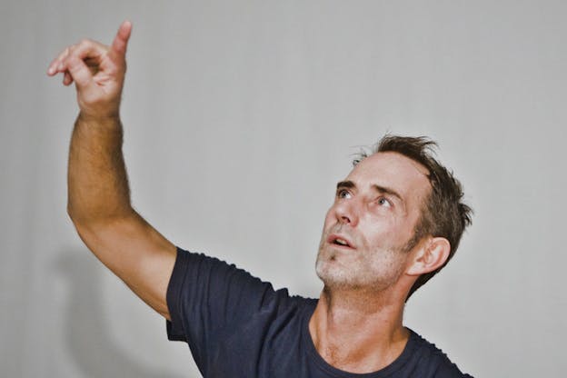 Close-up of a person in a dark gray tee-shirt holding up their arm and pointing their index finger against a light gray background.
