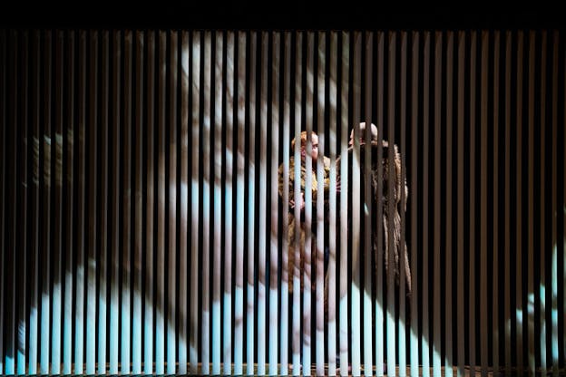 Two performers in fur coats face each other, their bodies partly obscured by a thin paneled screen displaying a close-up projection of a woman's face. 