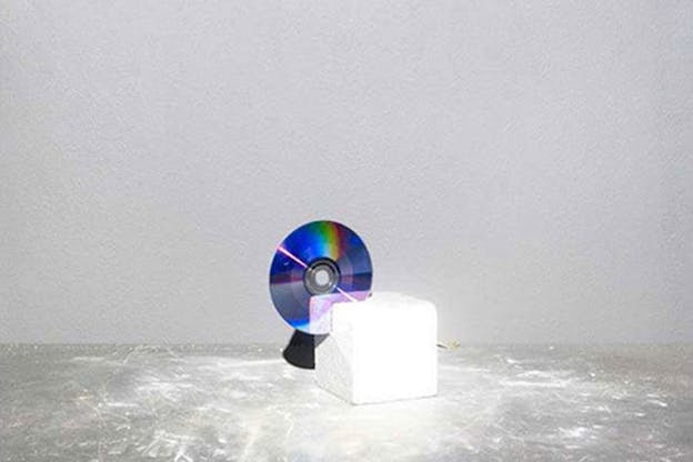Still-life of an iridescent CD wedged into a white block against a light gray wall. 