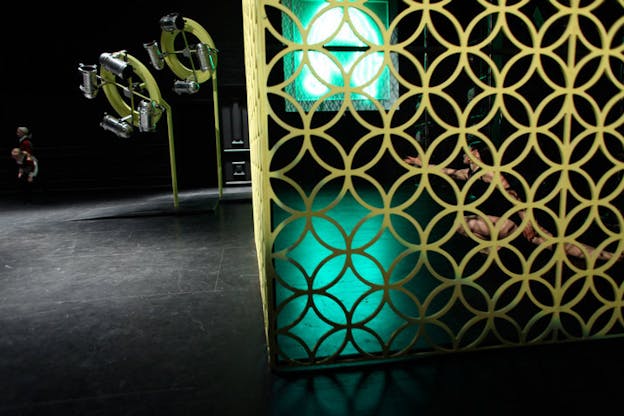 A performer wearing a black leotard and gold hoop earrings lunges within a geometrically patterned cage. Within the cage, there is also a green neon outline of the head of a person with bangs and long hair. Outside of the cage, there are two green structures composed of a rod connected to a circle at top and adorned with silver mechanical objects. Beyond these structures, there are two performers, dressed in red.