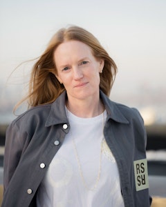 Anne Collier with light red hair swept back from the wind and blue eyes, wears a white shirt underneath a gray jacket with silver button on the right side and on the left a patch with the letter “RS” mirrored. A gold delicate chain necklace sits on top of the shirt. 