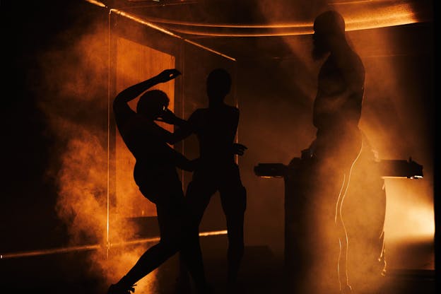 Three performers dance on a dark foggy stage, backlit by orange light. Two dancers careen as they embrace each other on the left side of the frame. The third stands straight in profile, facing them.