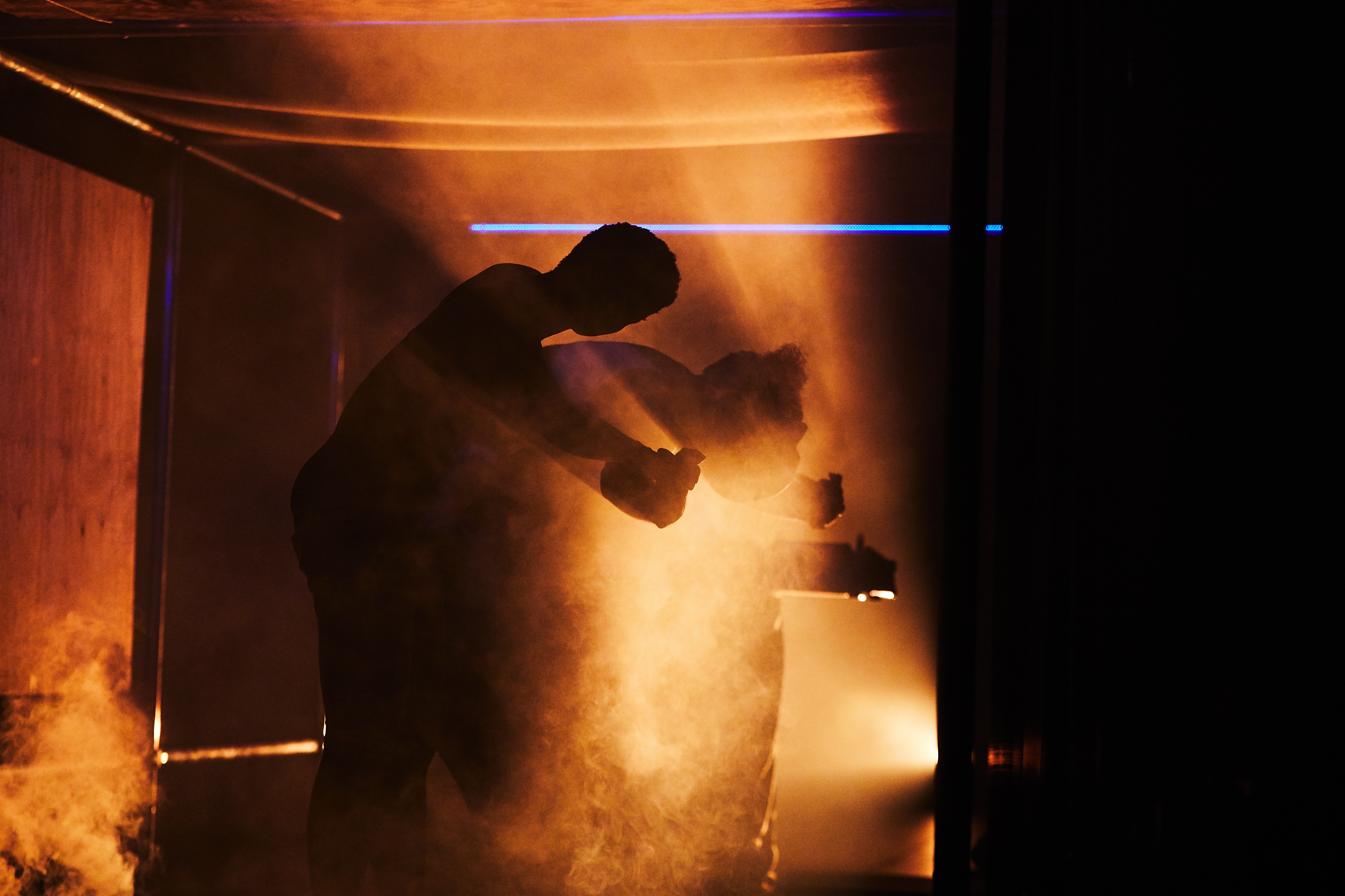 Two performers dance on a foggy stage, backlit by orange light. They stand facing each other, the dancer on the right arching backwards and the dancer on the left leaning over them as they do so.
