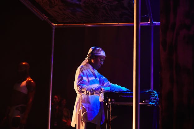 Yulan Grant performs on a stage within a metal frame, lit by blue and red light. The are standing in profile facing the right, adjusting the dials on a sound board in front of them. They wear black headphones over their ears, eyeglasses, a white durag, and a white button up shirt.