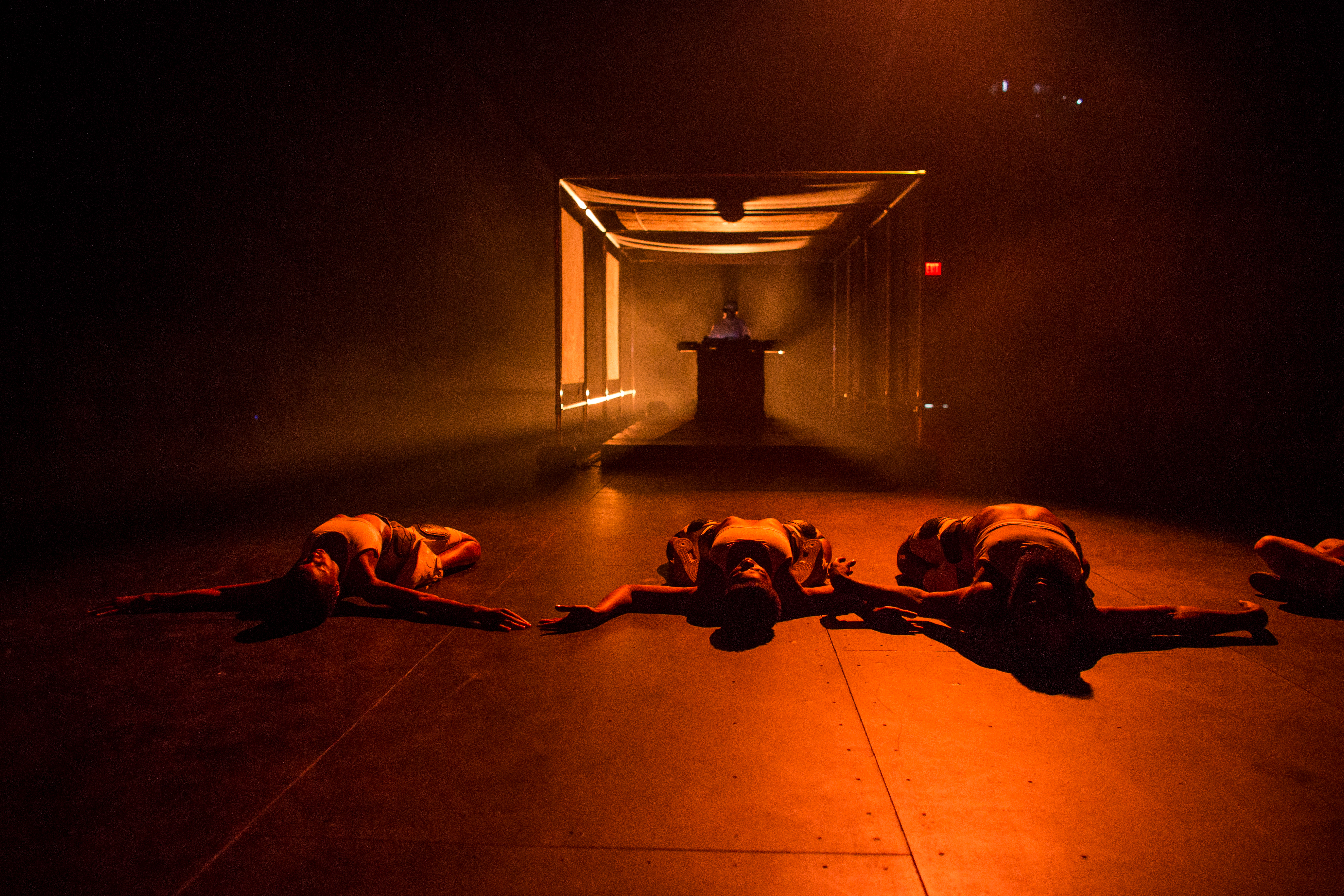 Three performers dance on a dark stage, lit with orange ambient light. They lay on their backs with their heads pointing towards the camera, arms spread, and legs folded under them. In the far background, Yulan Grant stands at a soundboard.