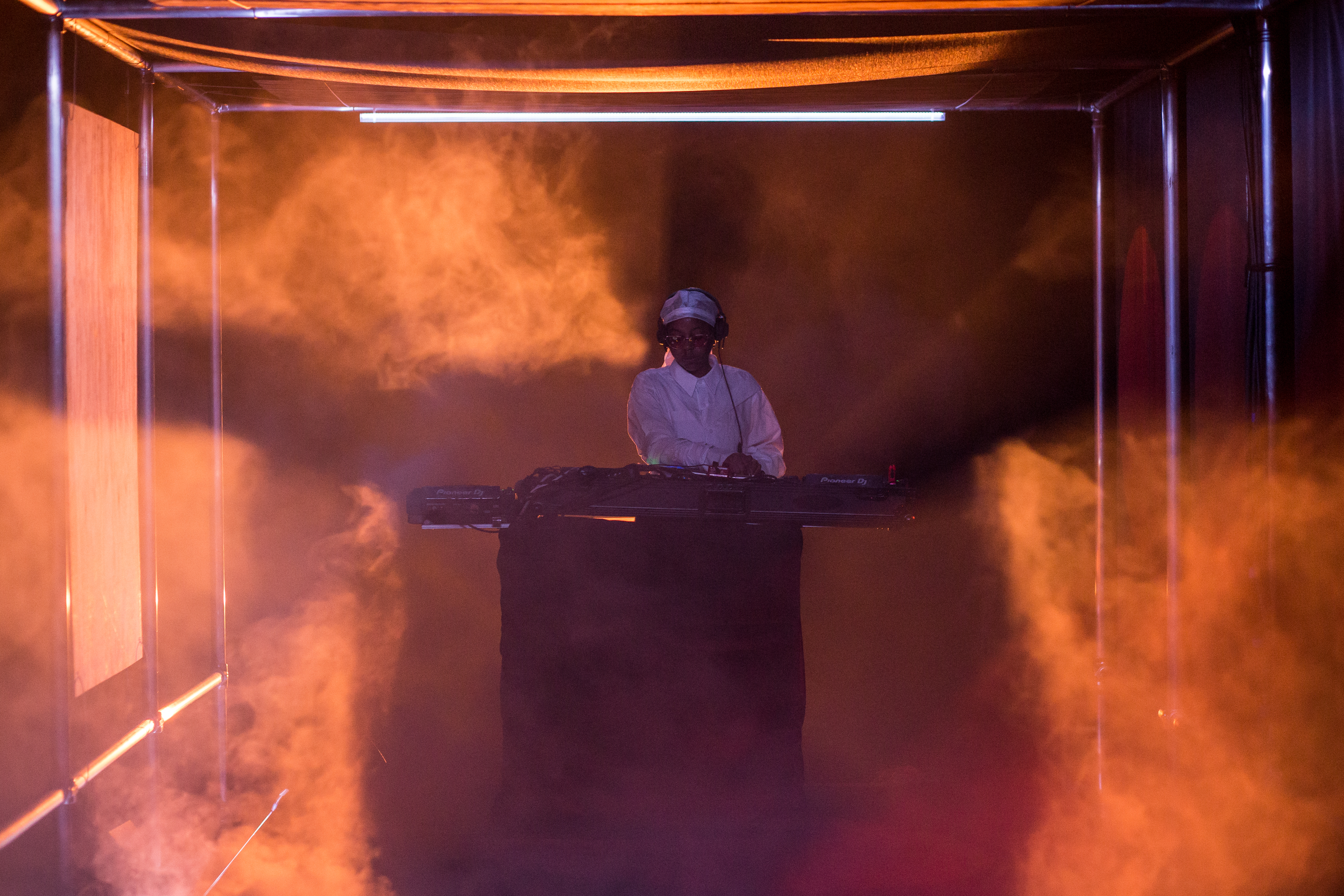 Yulan Grant performs on a foggy stage, backlit by orange light. They stand behind a podium with a soundboard, dressed in a white du-rag and white button up shirt. 