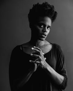 Portrait of Okwui in black and white. She stands, with her hands raised in front of her chest, loosely joined together. Her head is tilted to the left but she looks directly into the camera.